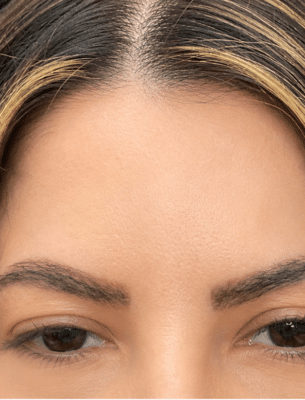 Close-up of the upper part of a face: smooth forehead, arched eyebrows, and brown eyes partially visible at the bottom of the frame, with dark hair and lighter highlights at the hairline.