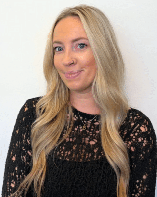 Portrait of a smiling blonde woman with blue eyes wearing a black lace top and a small nose hoop, facing slightly left with a white background.