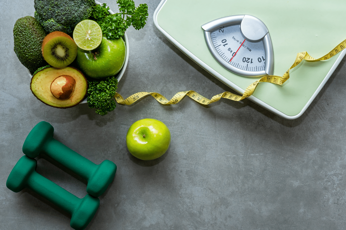 Green kitchen scale with a measuring tape and a bowl of fresh fruit and greens, suggesting healthy weight management.