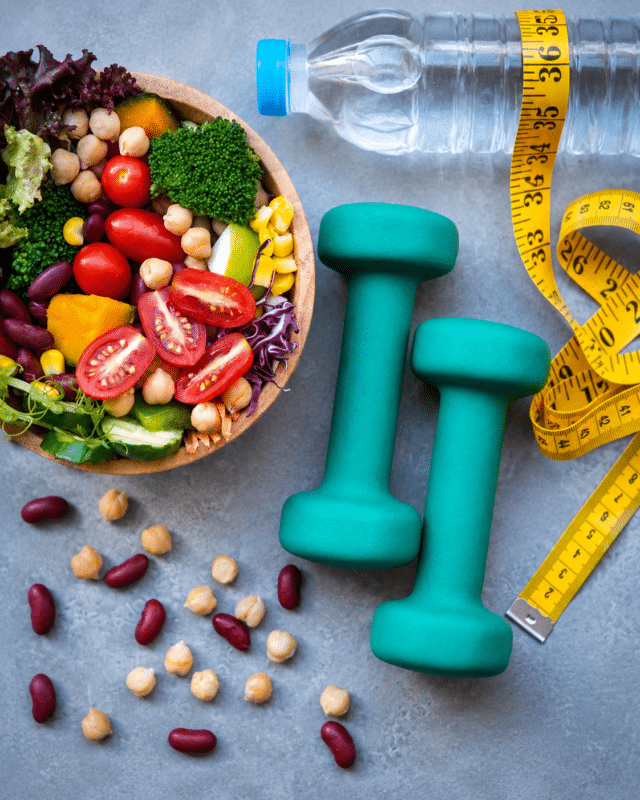 Colorful vegetable salad in a bowl beside turquoise dumbbells, a yellow measuring tape, and a water bottle—fitness and healthy eating setup.