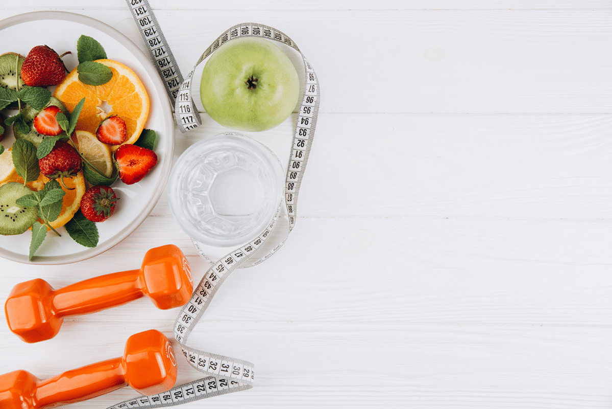 Plate of sliced citrus, berries, kiwi and mint with a green apple, water glass and measuring tape on a white surface, plus orange dumbbells—healthy living theme